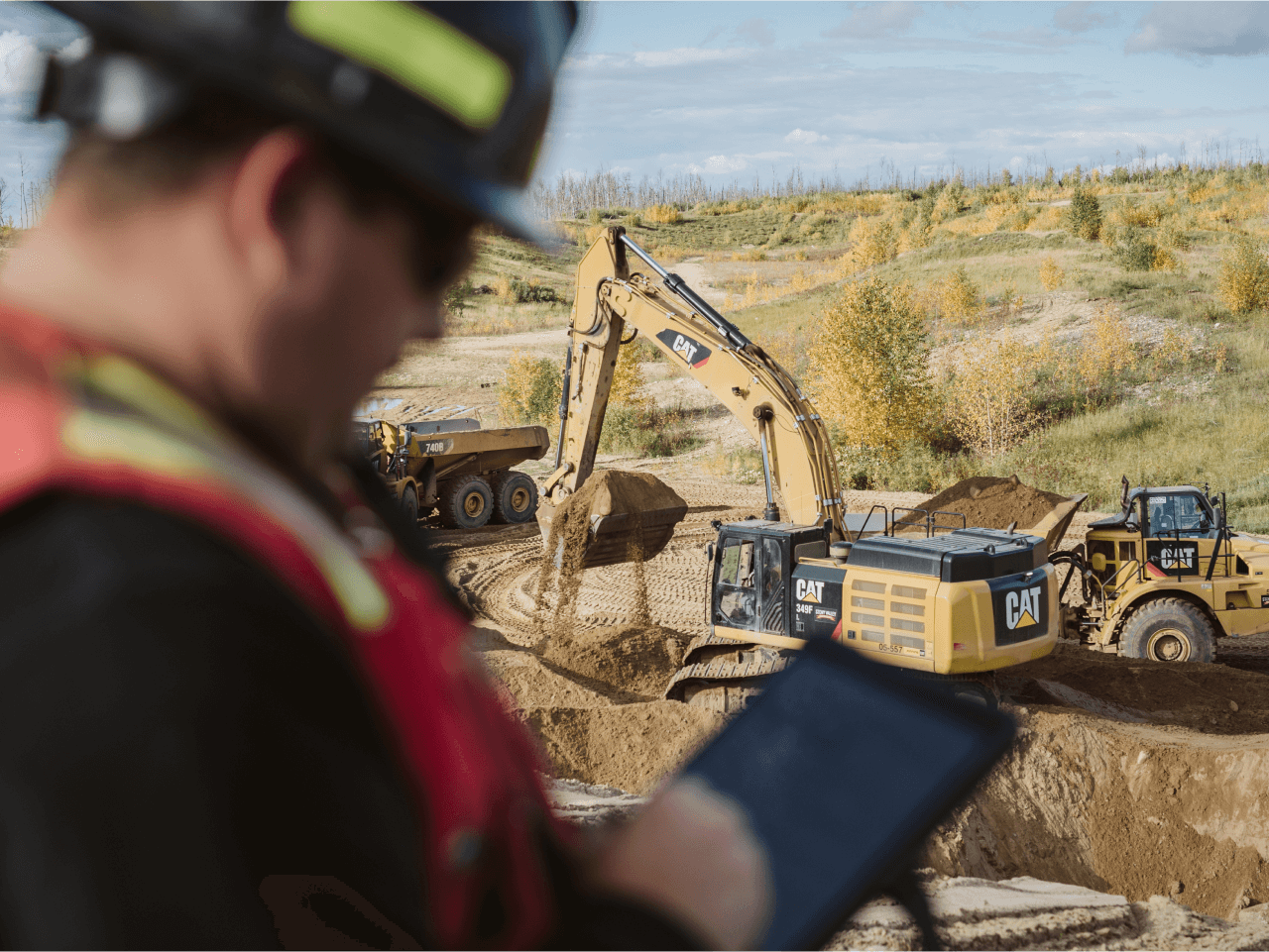 Field service worker using a tablet in the foreground while CAT heavy machinery operates in the background on a construction site.